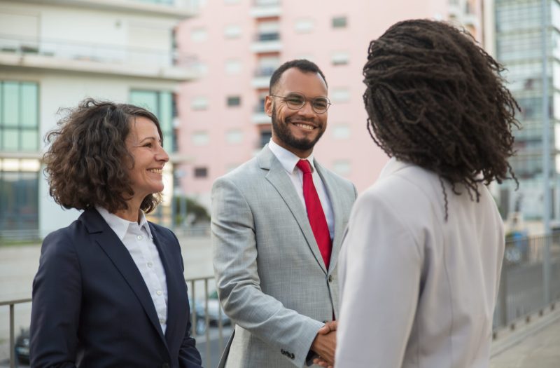 Agent meeting clients near office building. Business man and women standing in city, shaking hands and smiling. Friendly talk concept